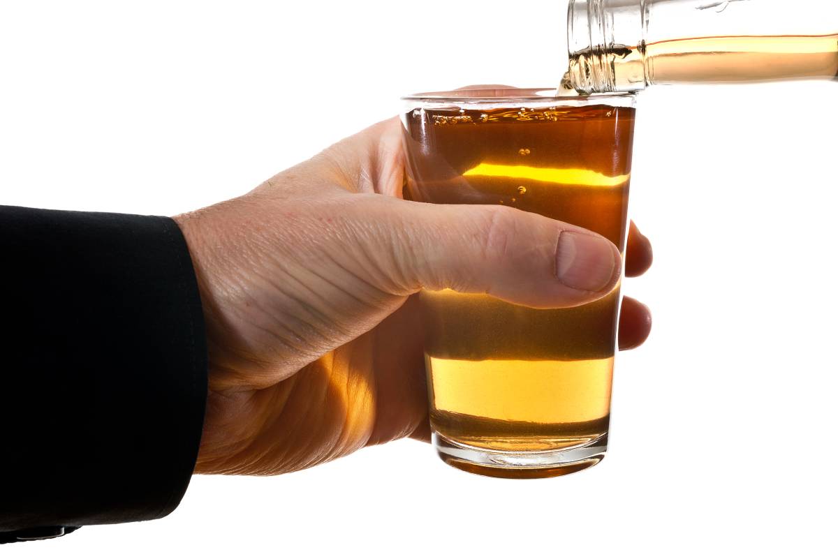 Billedet viser en hånd holder et glas med en gylden væske, mens en flaske hælder mere af den samme drik i glasset.